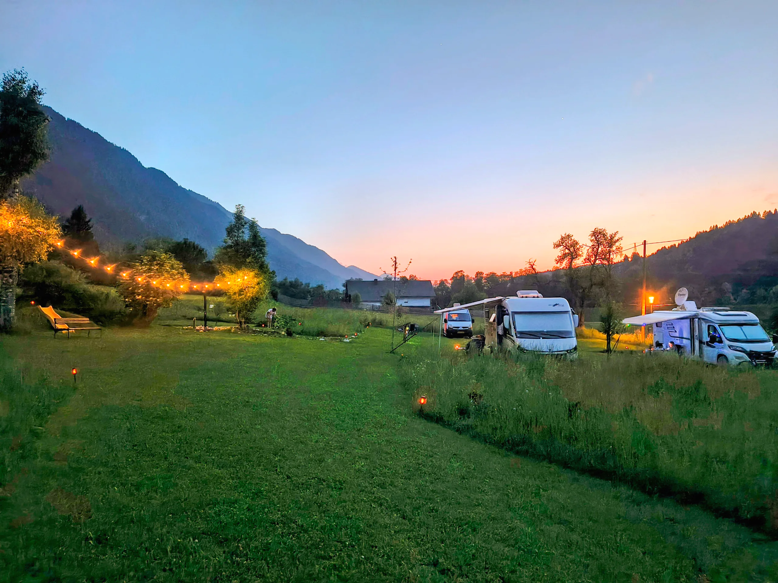 Wohnmobile auf der Stellplatzwiese, während der Abendhimmel hinter den Bergen orange wird