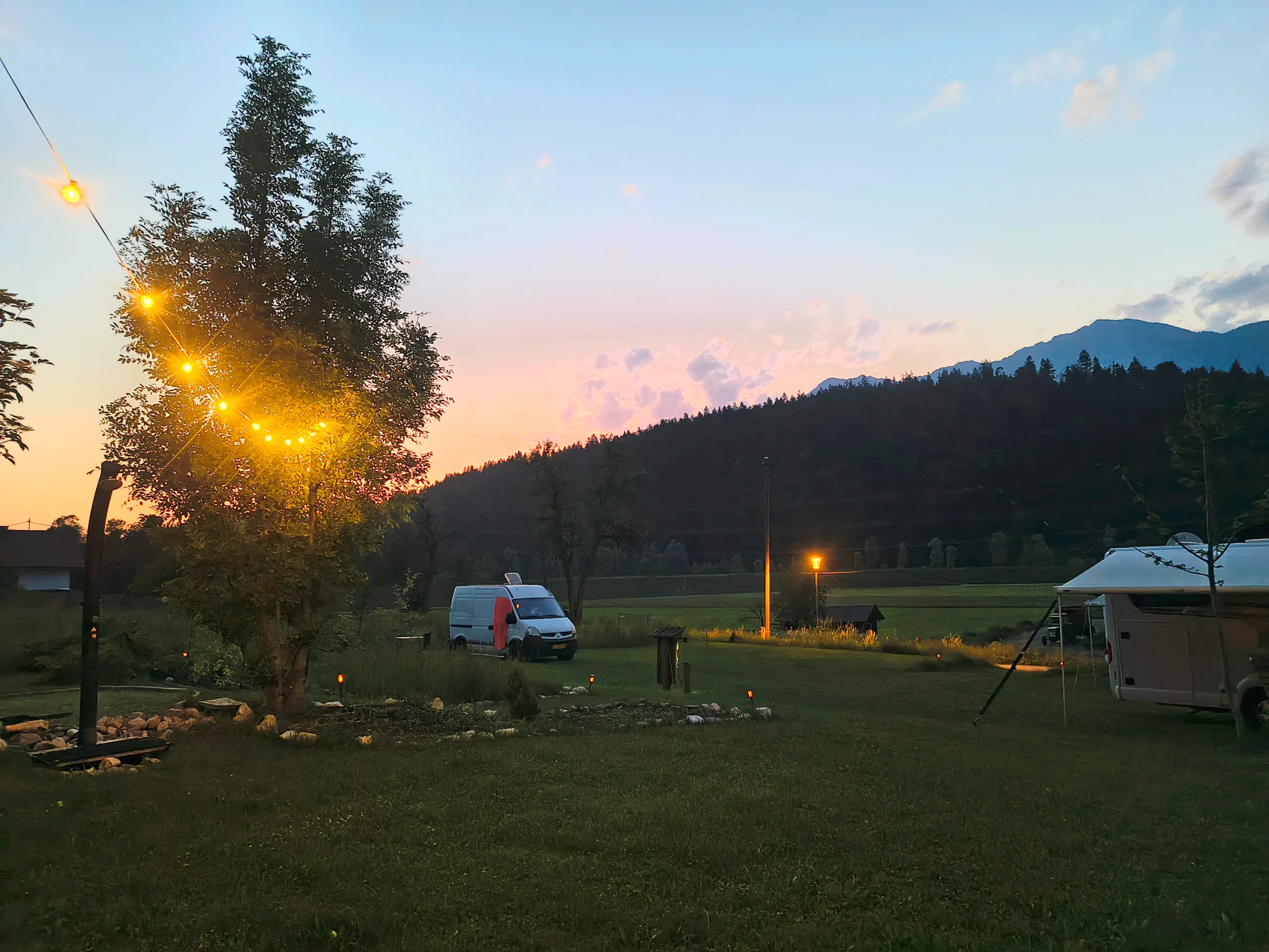 Lichterkette im Baum über der Stellplatzwiese, dahinter ein Campervan und der rosa Abendhimmel über dem Waldrücken