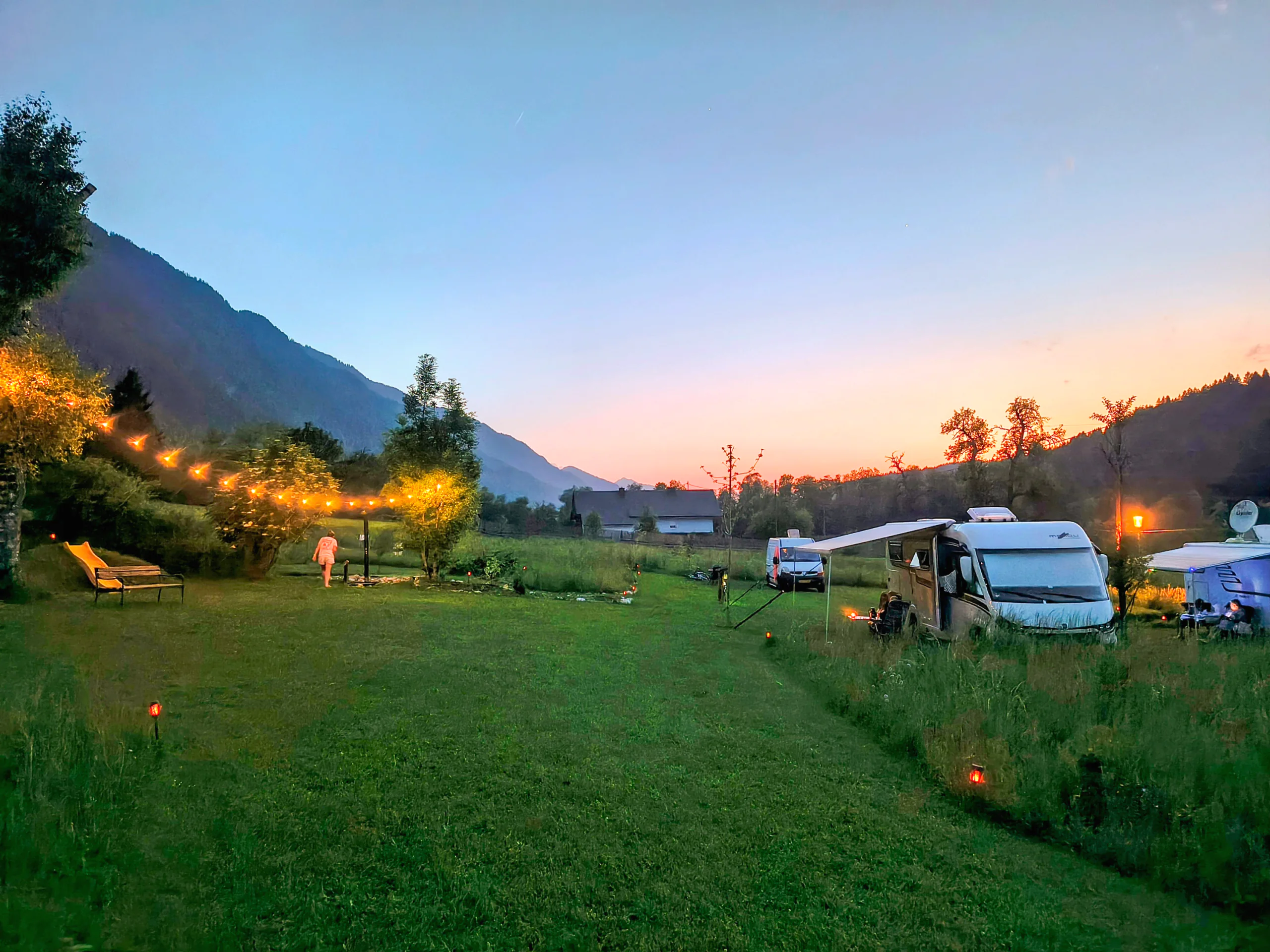 Stellplatzwiese in der Dämmerung, eine Lichterkette spannt sich zwischen die Bäume, am Rand stehen Wohnmobile