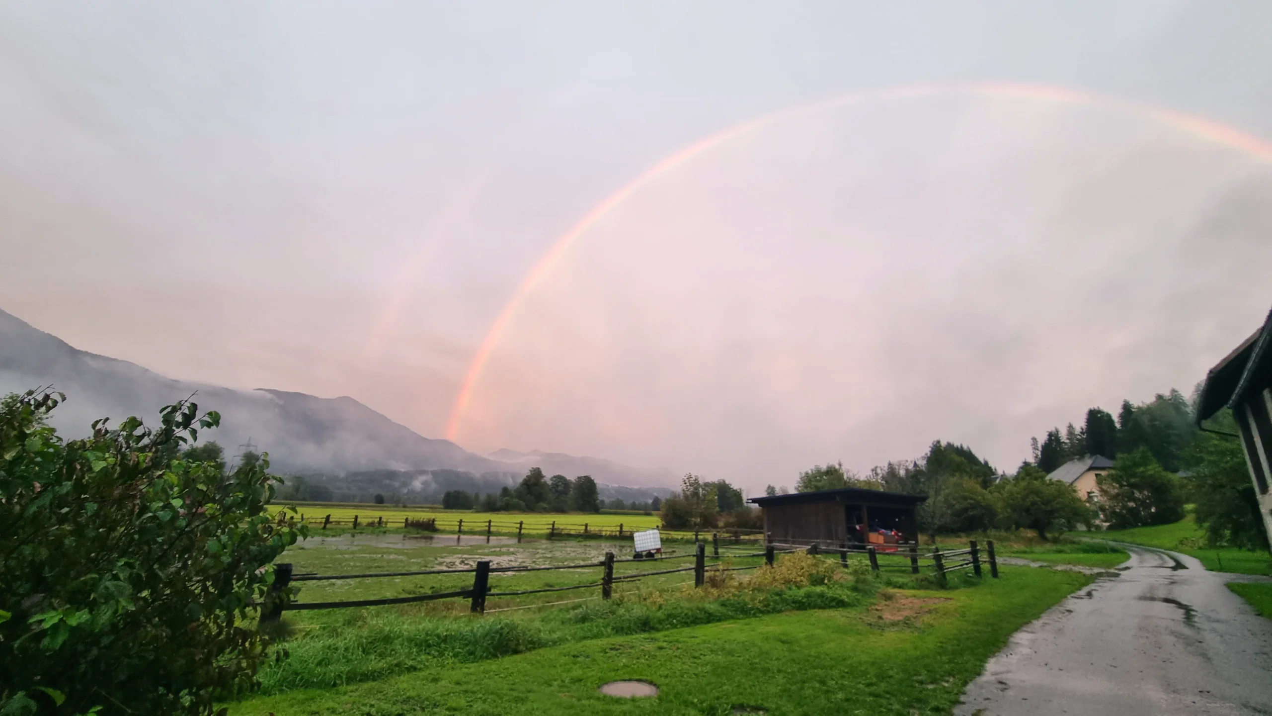 Regenbogen über dem Tal nach einem Regenschauer, im Vordergrund der nasse Weg am Zaun