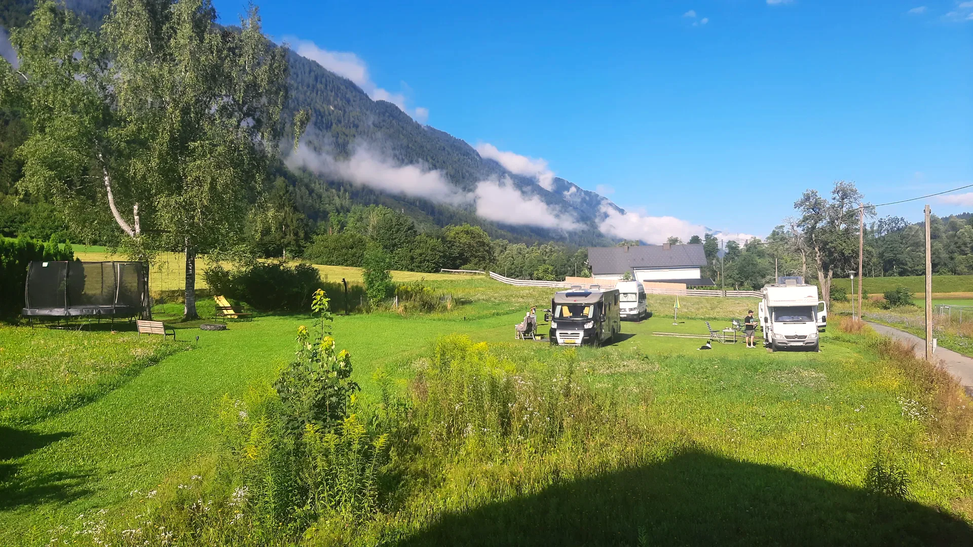 Wohnmobile auf der Stellplatzwiese, tief hängende Wolken über dem Berghang, links das Trampolin