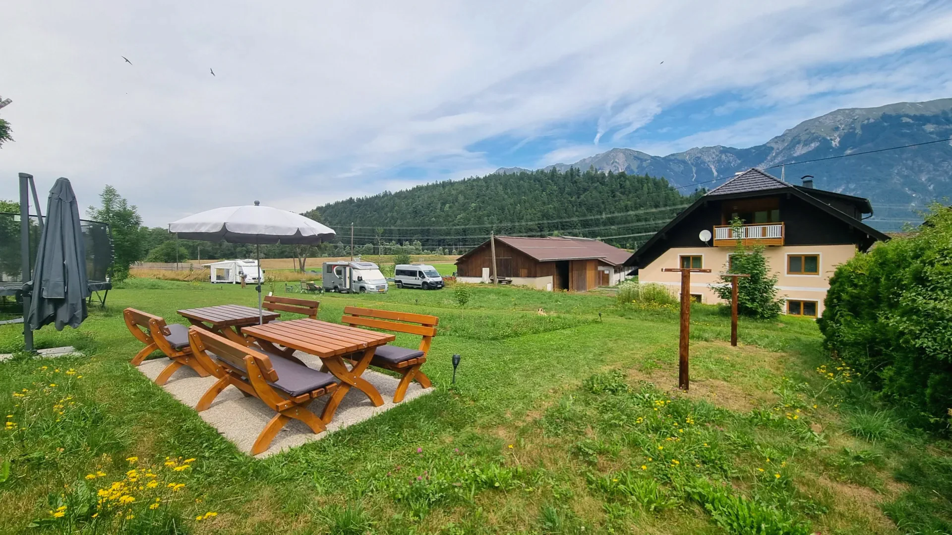 Sitzgruppe unter dem Sonnenschirm auf der Wiese, dahinter die Wohnmobile, der Stadel und das Haus vor den Bergen