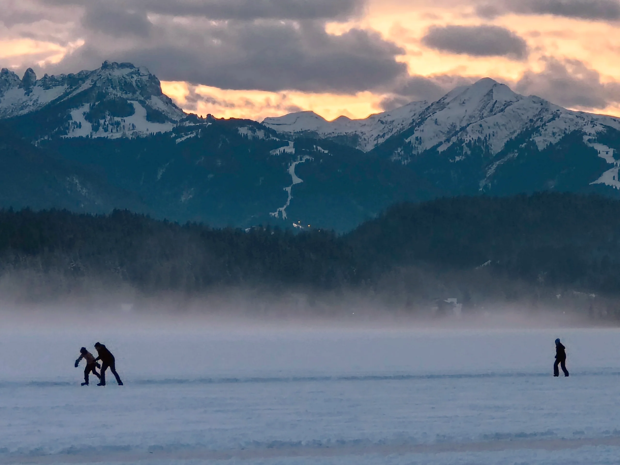 Eisläufer auf der weiten Natureisfläche im Abendlicht, über dem Nebel stehen die verschneiten Berge