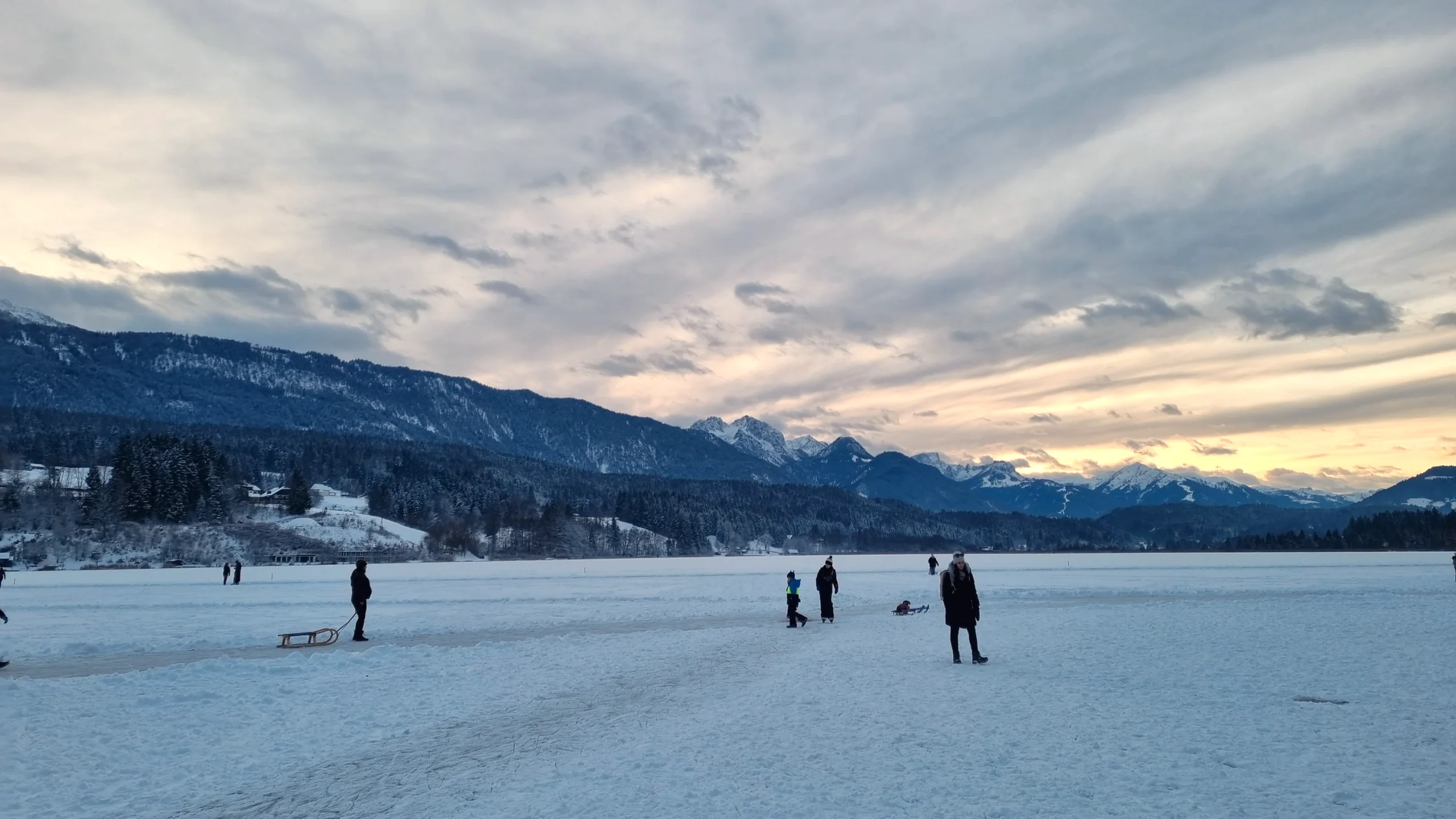 Menschen mit Schlitten auf der Eisfläche, über den Bergen steht ein heller Abendhimmel