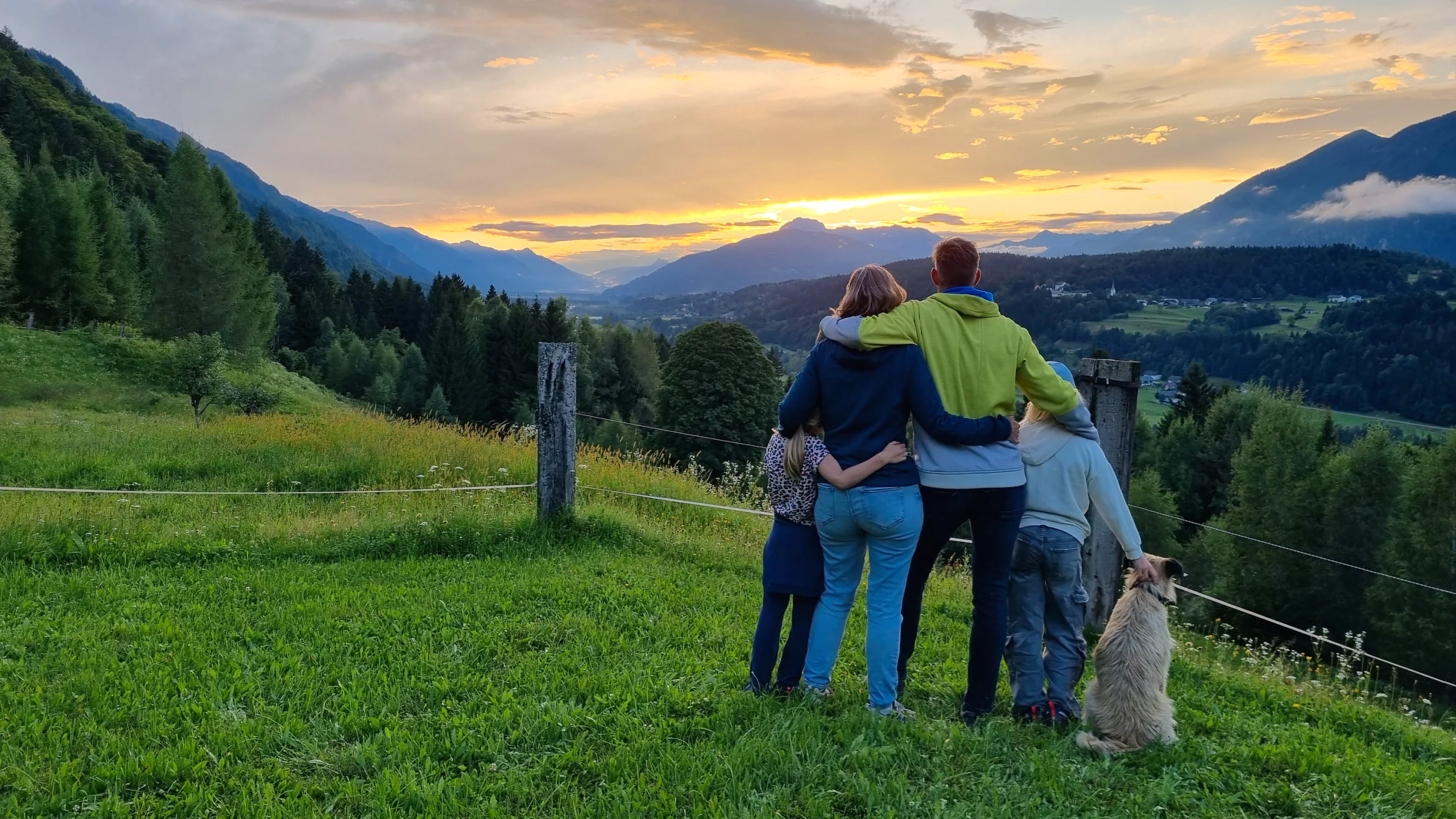Die Familie steht Arm in Arm mit dem Hund auf der Wiese und schaut in den Sonnenuntergang über dem Tal