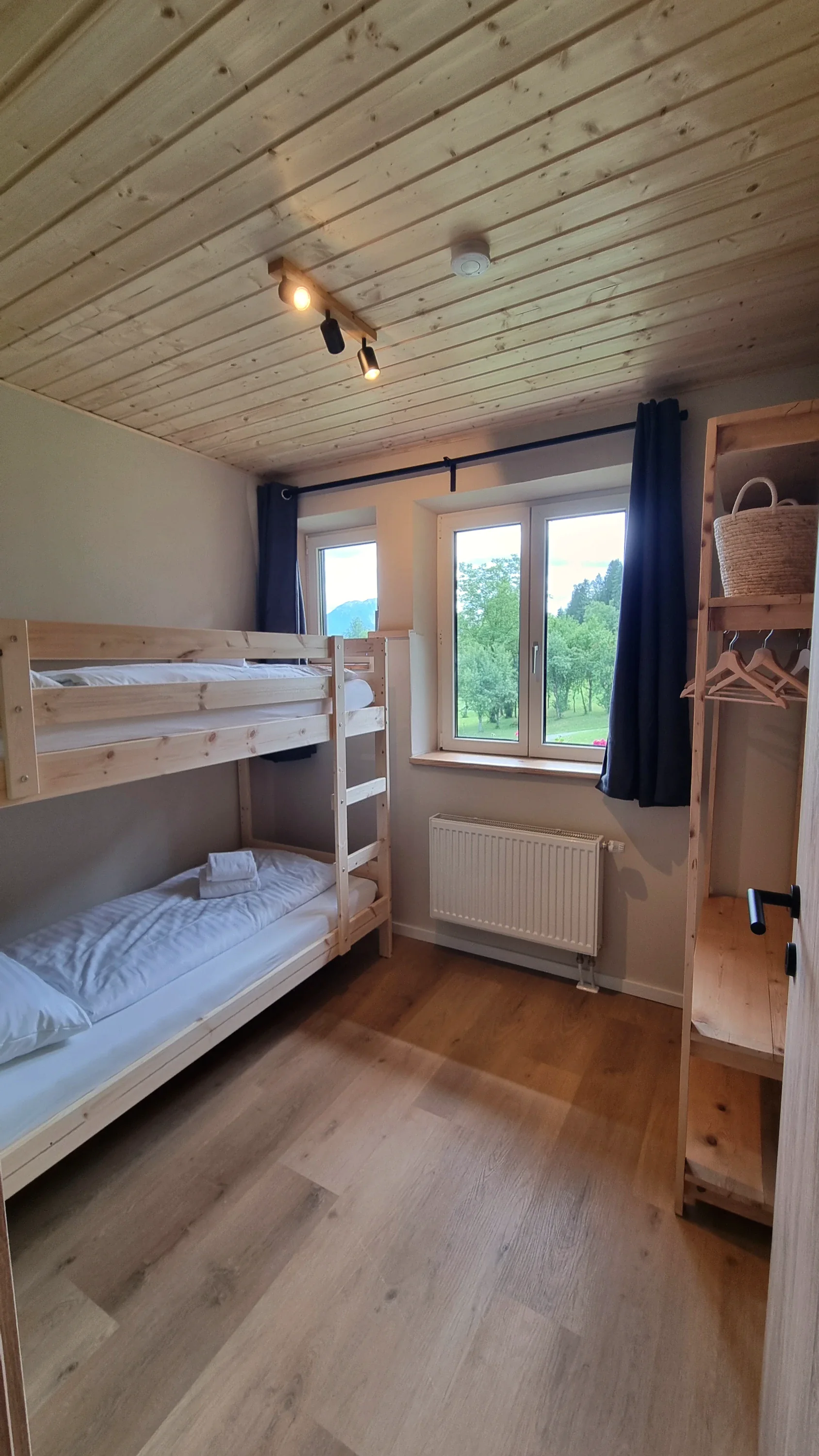 Stockbett aus hellem Holz am Fenster, daneben ein offenes Regal, draußen das Grün