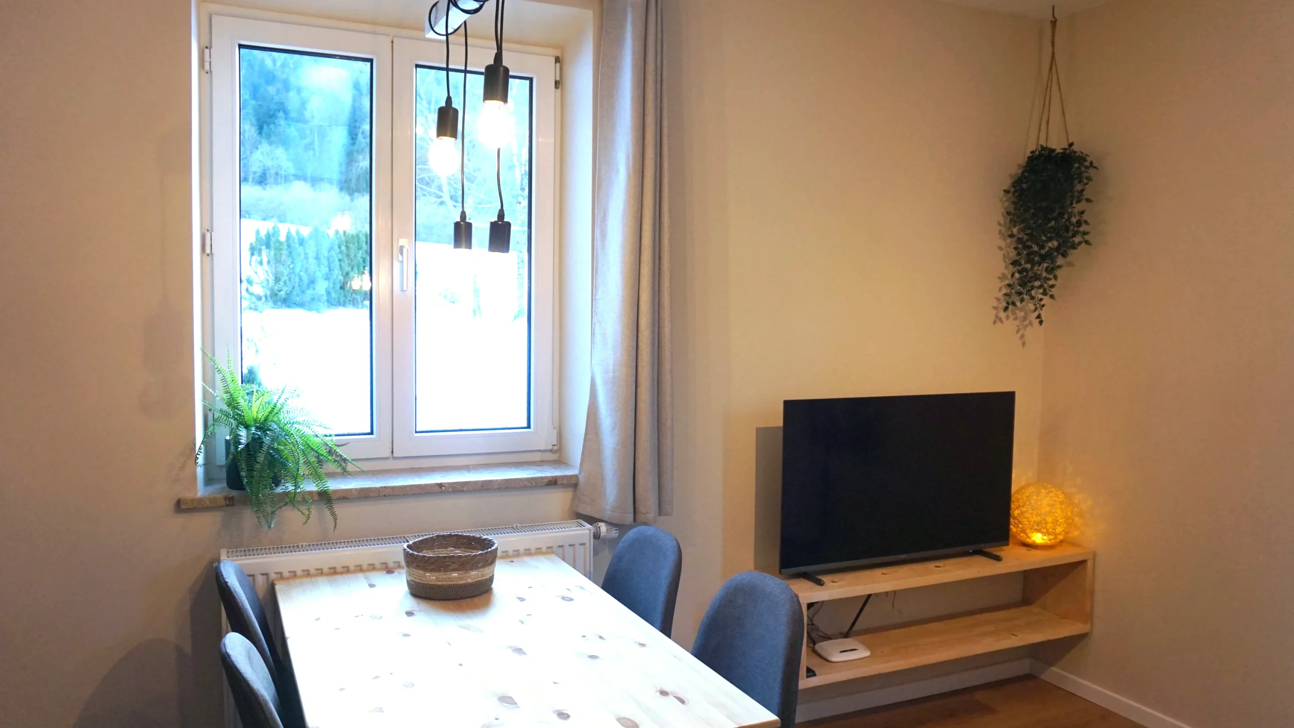 Wohnzimmer der Ferienwohnung 1 mit Esstisch, Fernseher auf dem Holzregal und Fenster zum verschneiten Garten