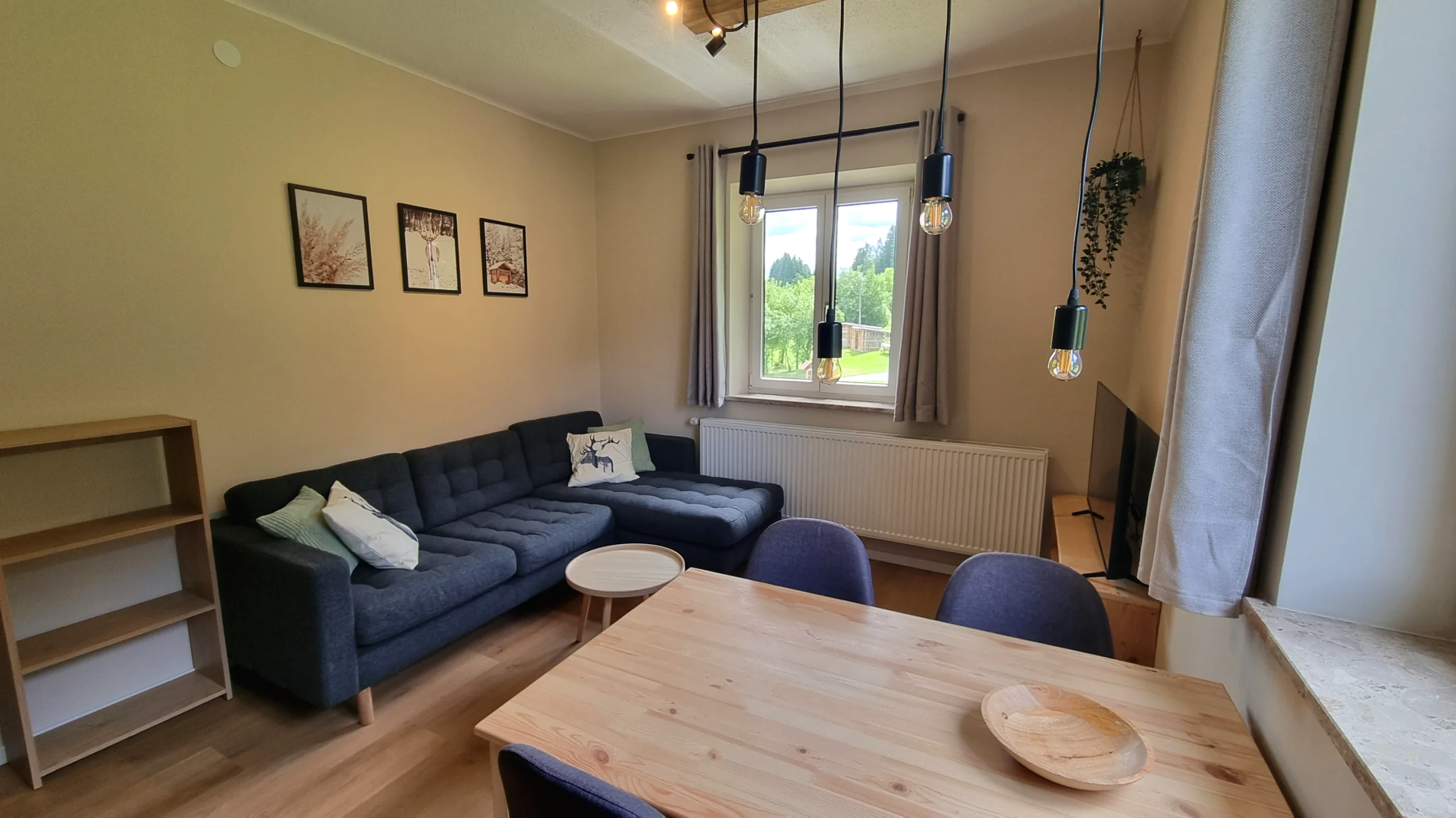 Wohnbereich der Ferienwohnung 2 mit dunkelblauem Ecksofa, Esstisch aus Holz und Pendelleuchten vor dem Fenster