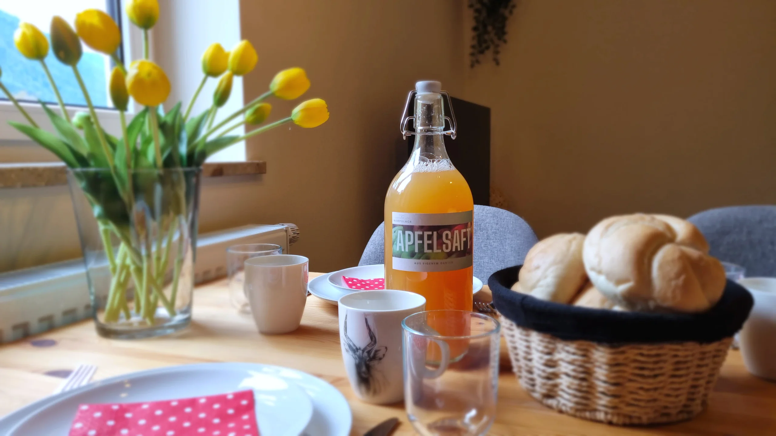 Frühstückstisch mit Brötchenkorb, Apfelsaft und Tulpen am Fenster