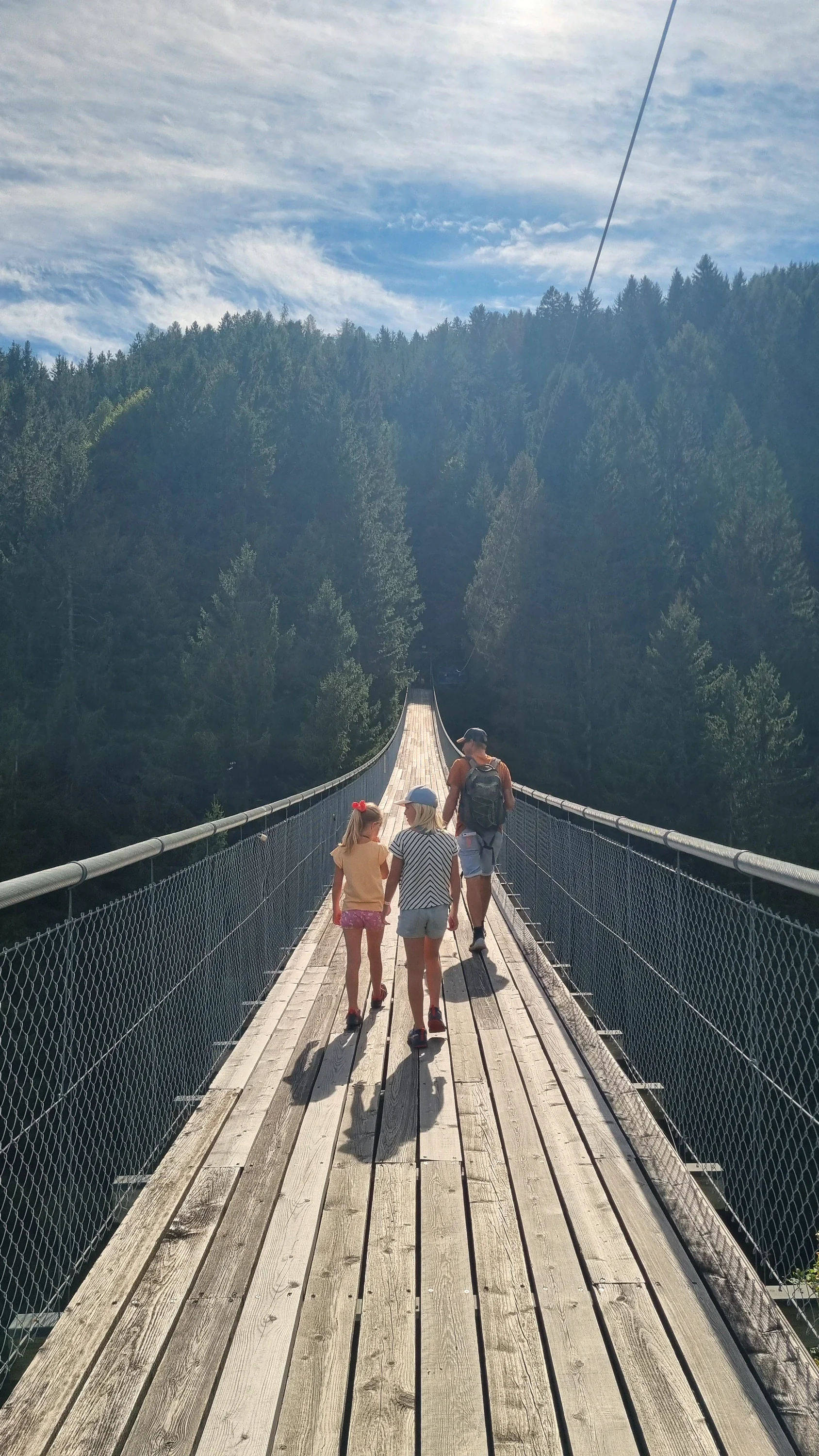 Eine Familie geht über eine Hängebrücke aus Holzbohlen, links und rechts fällt der Wald ab