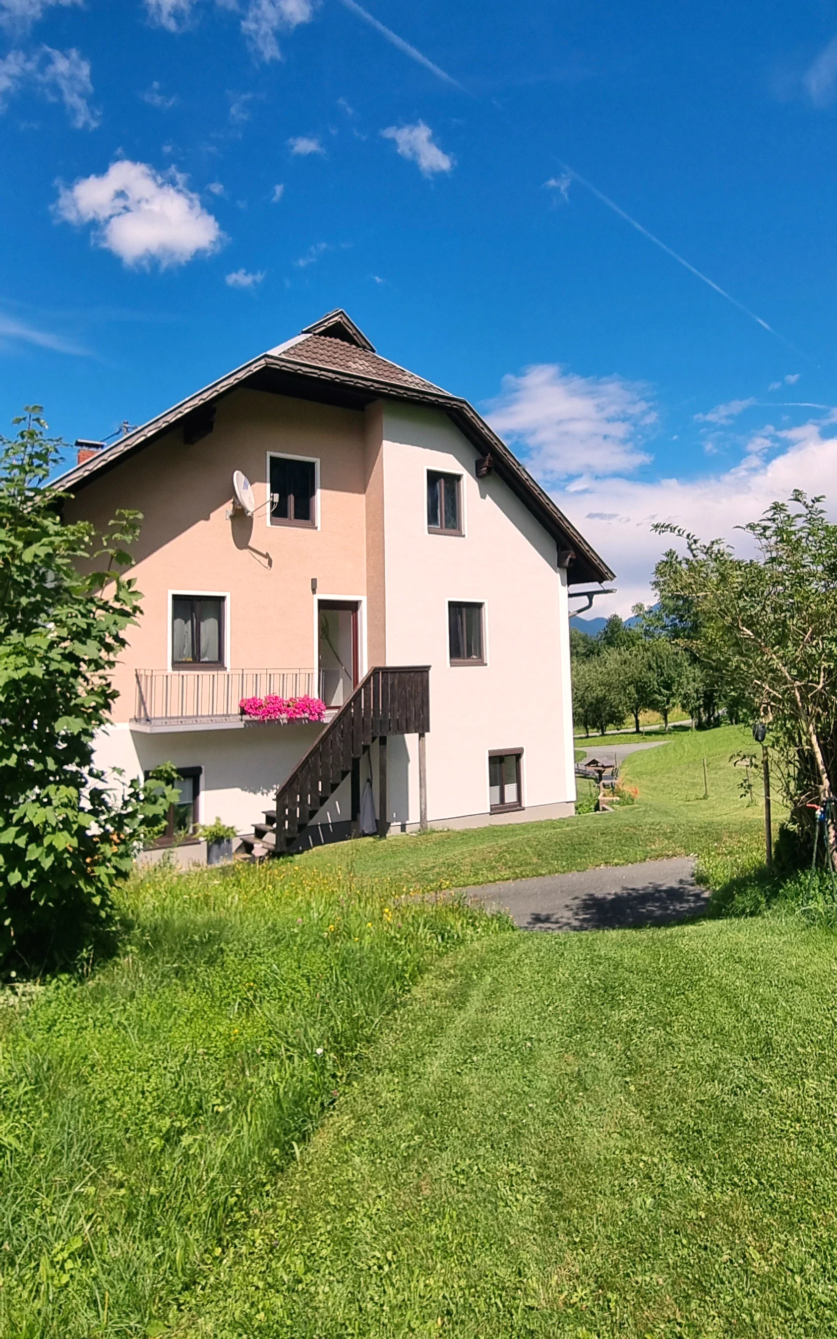 Haus Nampolach im Sommer unter blauem Himmel, die Wiese führt zum Eingang, Geranien leuchten am Balkon
