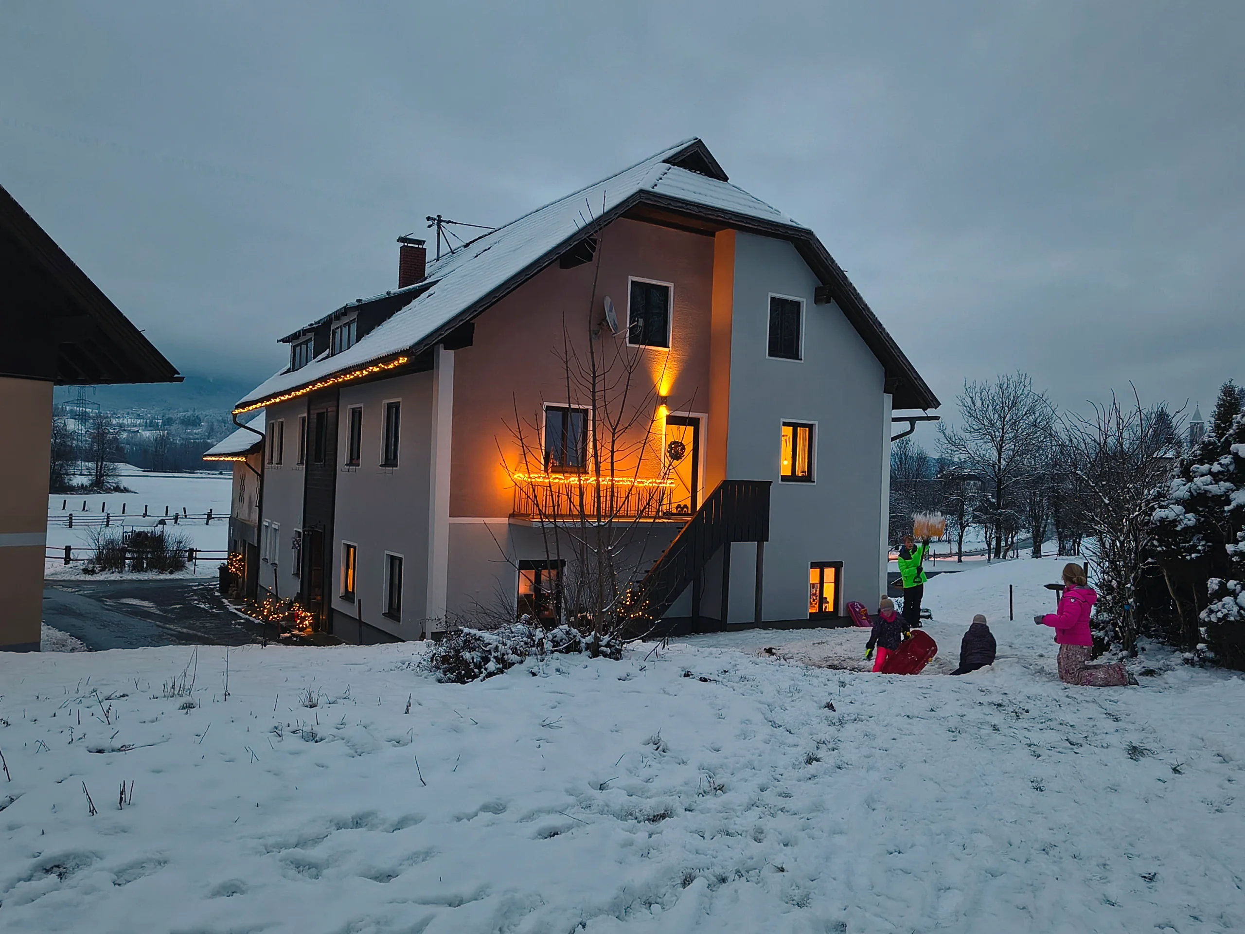 Haus Nampolach zur blauen Stunde im Schnee, die Fenster warm erleuchtet, Lichterketten am Dach, Kinder rodeln davor