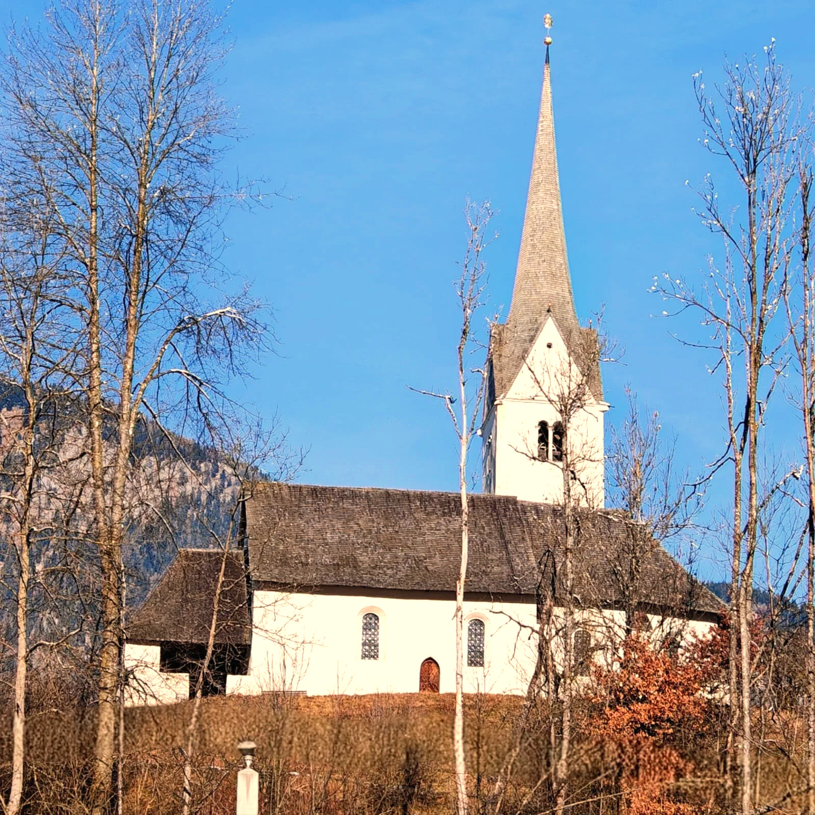 Kleine Kirche mit spitzem Turm und Schindeldach im Schnee, ringsum kahle Bäume