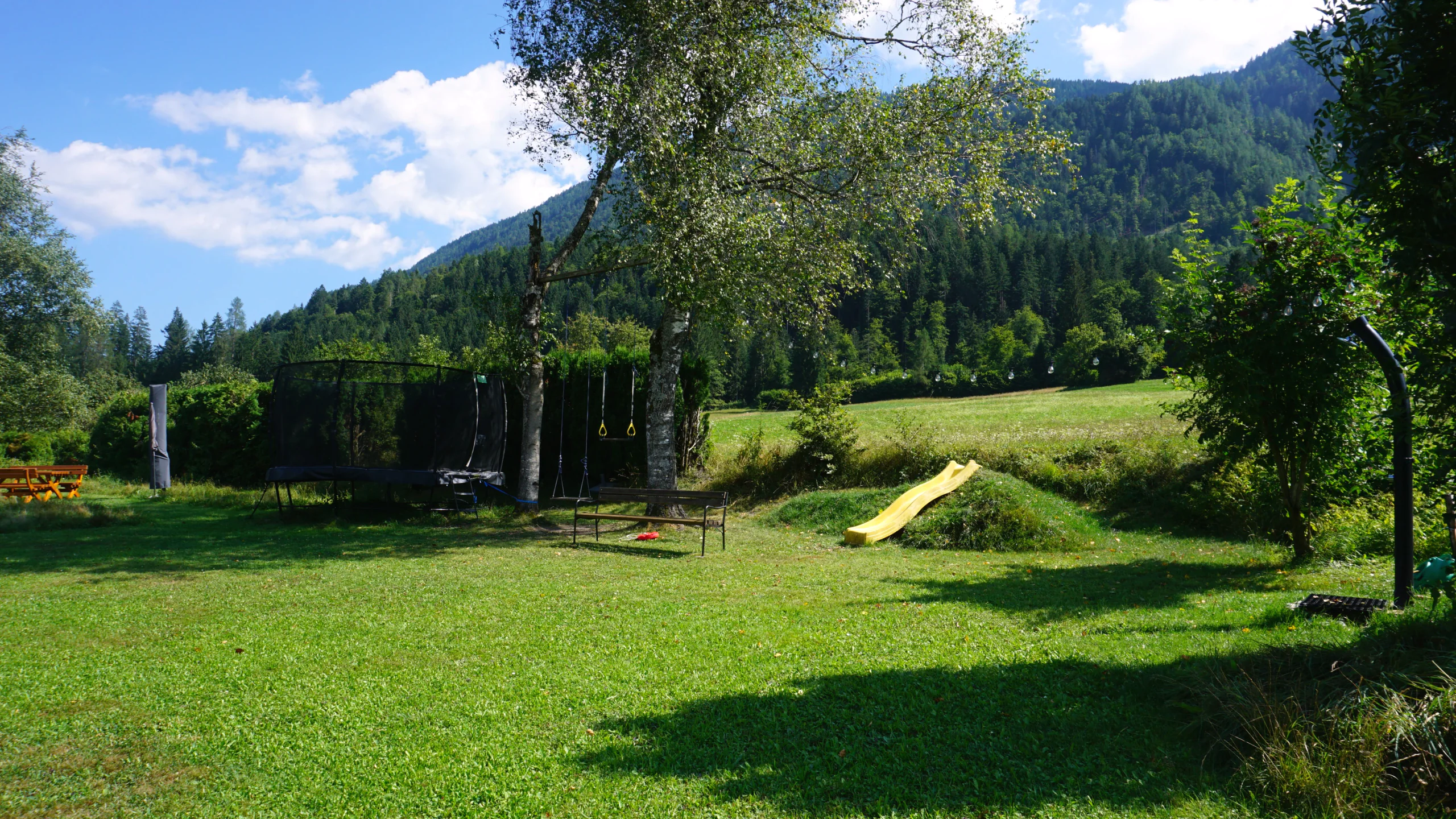 Wiese mit Trampolin, Schaukel und gelber Rutsche, dahinter bewaldete Berghänge
