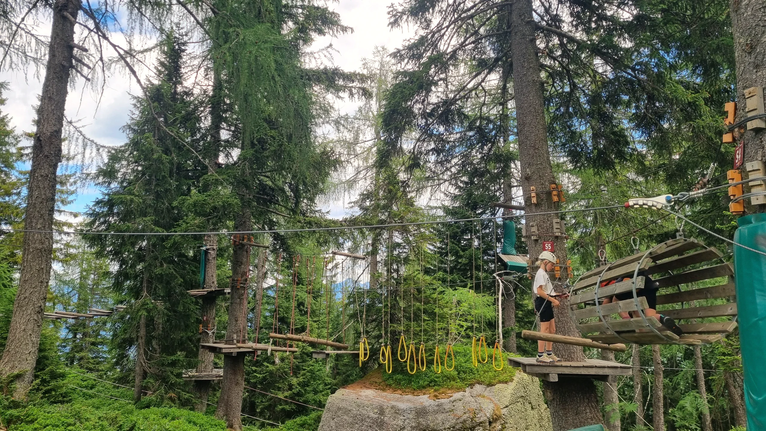 Kletterpark im Wald: Seilbrücken und Plattformen zwischen den Bäumen, ein Kind hängt gesichert an der Seilbahn