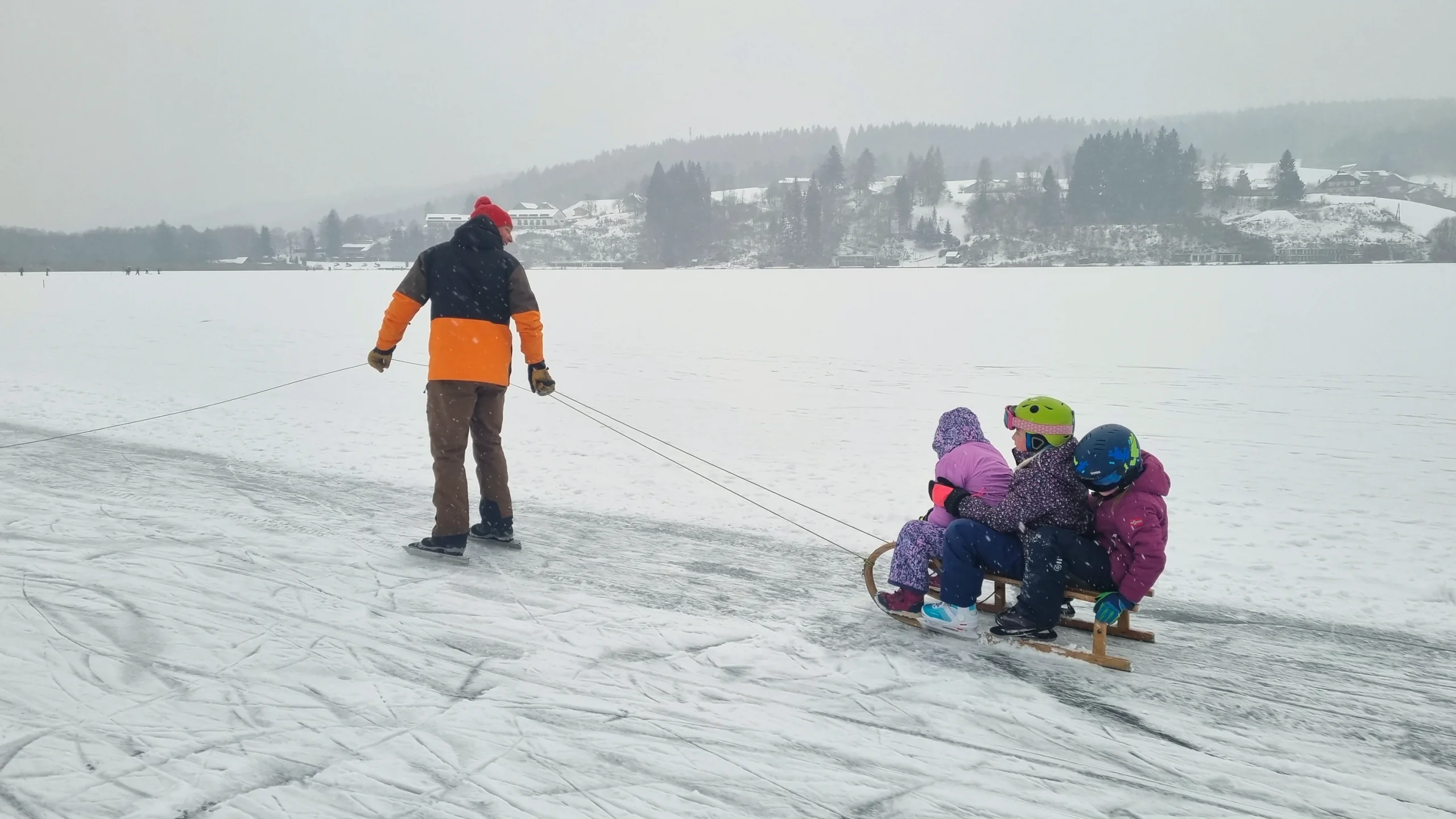 Ein Erwachsener zieht drei Kinder auf einem Schlitten über die Eisfläche, am Ufer liegt das verschneite Dorf