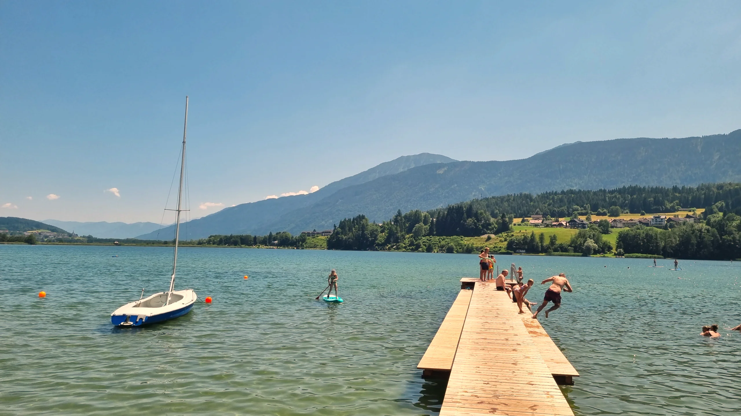 Holzsteg am Pressegger See mit einem Segelboot auf dem türkisen Wasser, dahinter bewaldete Berge