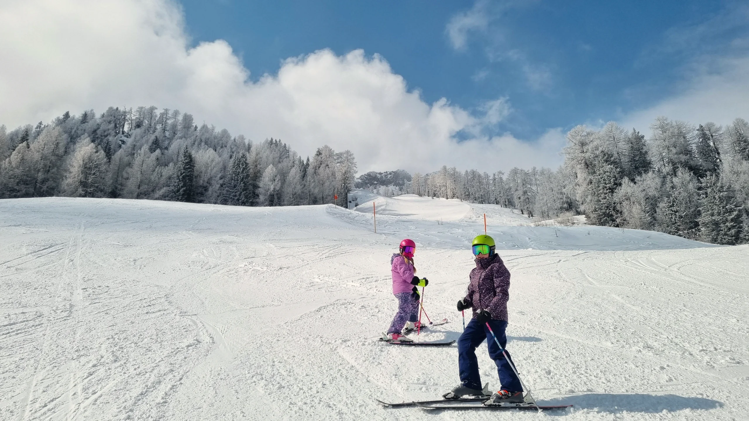 Zwei Kinder auf Skiern auf einer sonnigen Piste, dahinter raureifweißer Wald