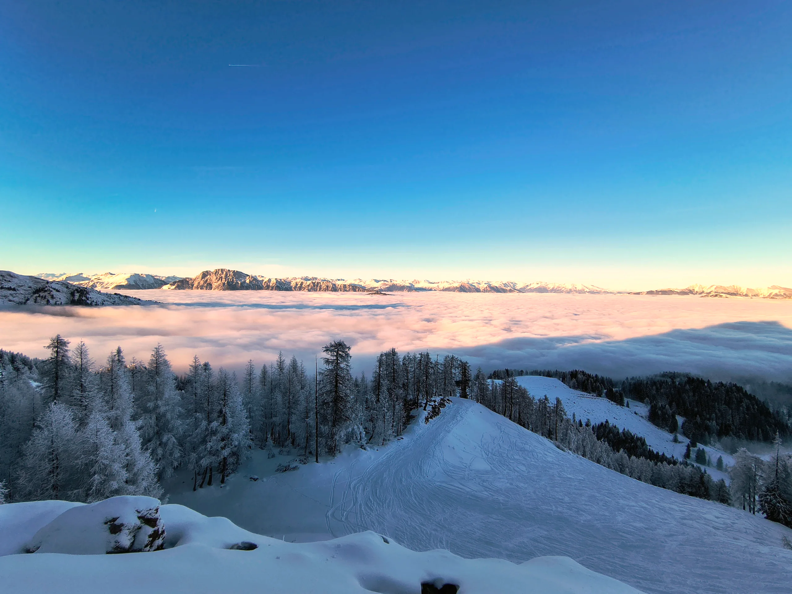 Verschneiter Hang über einem Wolkenmeer, am Horizont eine Kette von Gipfeln